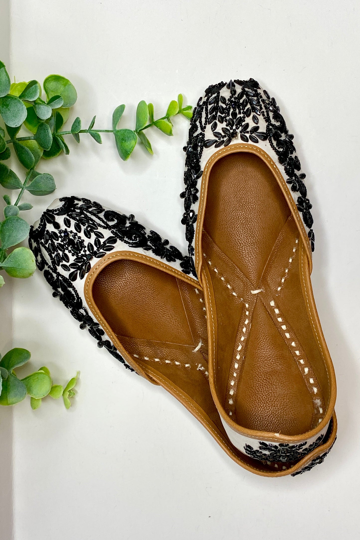 A pair of Nargis Jutti flats with black and white floral pattern on a flat surface with green leaves in the background.