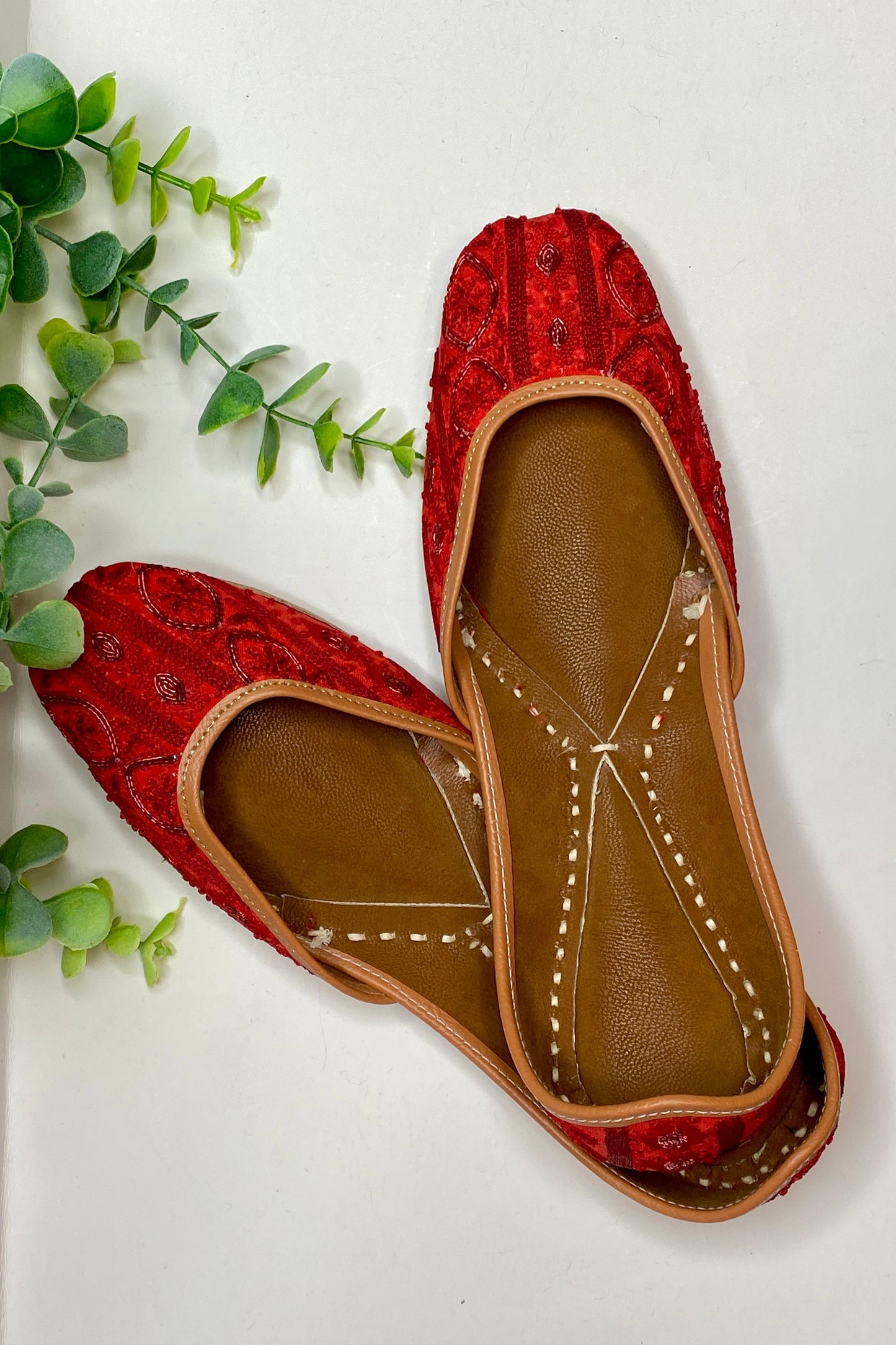 A pair of red and brown Cindy Jutti flat shoes with floral patterns and silver embellishments, displayed against a white background with green foliage to the side.