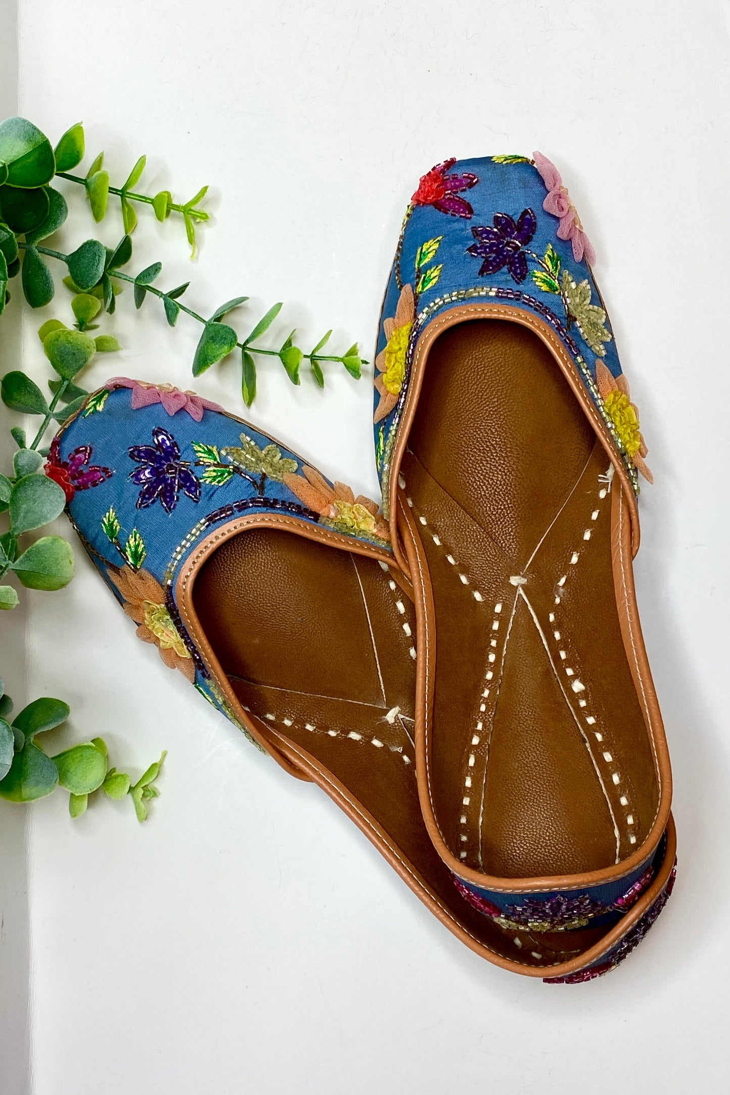 A pair of Gulabo Jutti shoes with floral patterns and a brown base color, displayed on a white surface with greenery in the background.