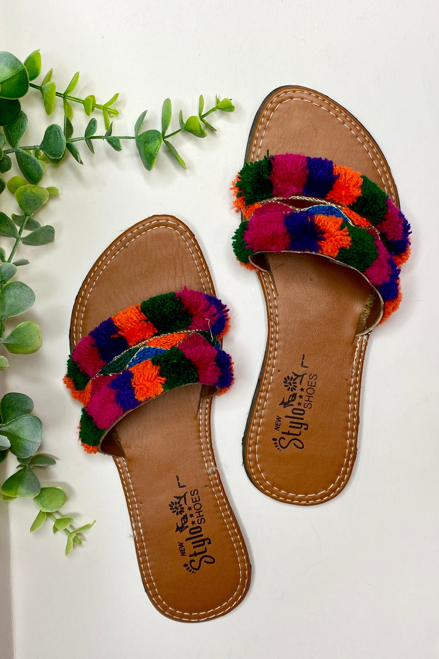 A pair of flat slip-on sandals with multicolored pom-pom straps on a white surface, with a green plant in the background.