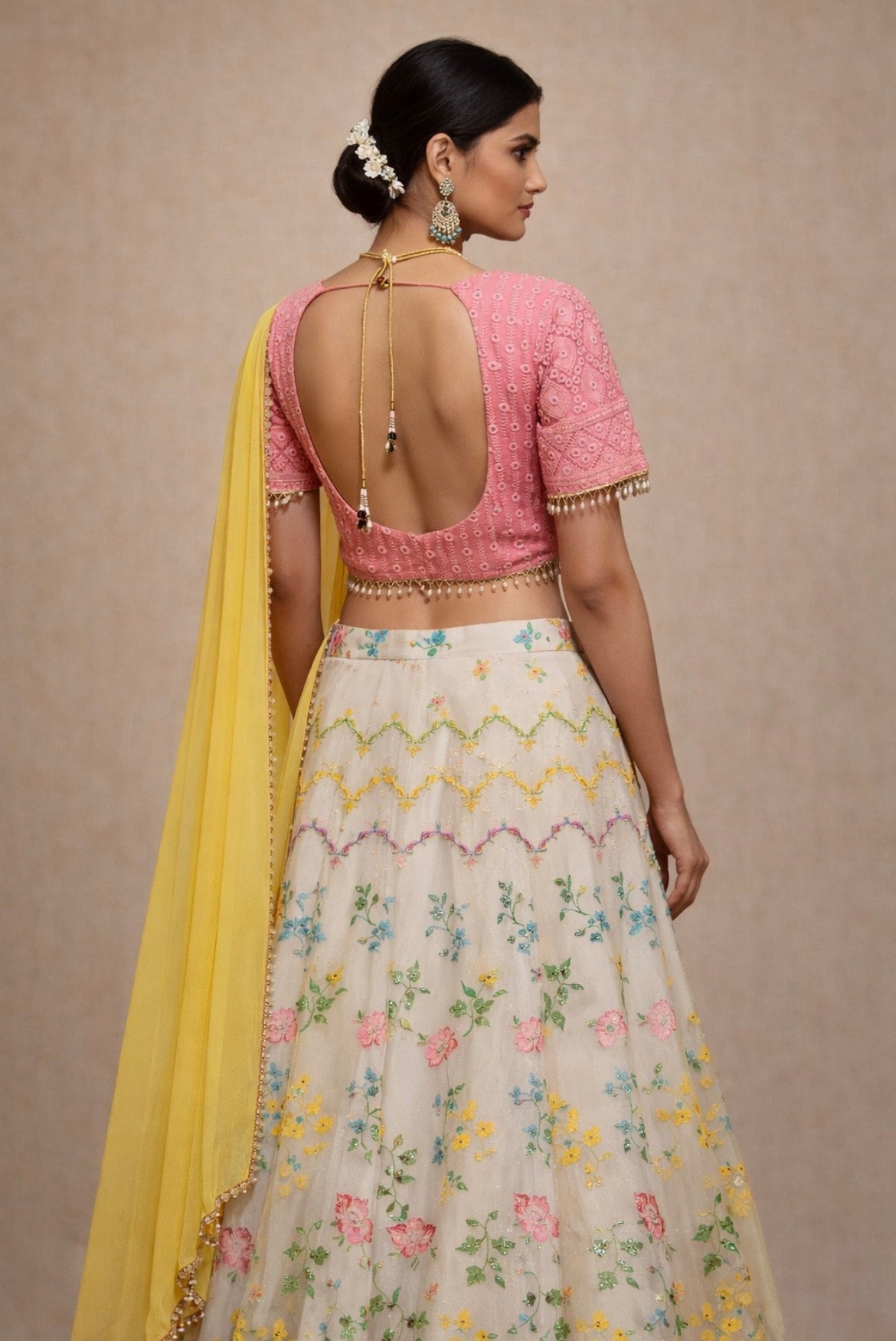 Woman wearing a pink blouse with an open back and a floral skirt with a yellow dupatta on a beige background