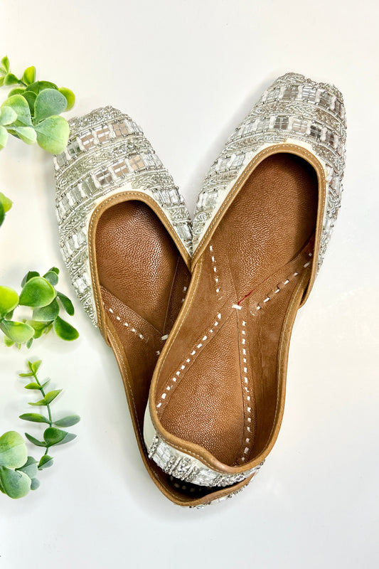 A pair of Sheesh Jutti shoes with silver embellishments on a white background, surrounded by green foliage.