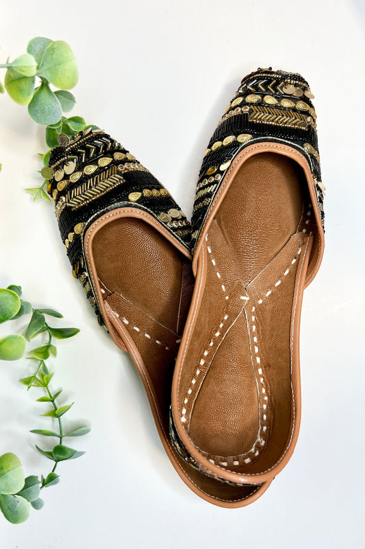 A pair of Urvashi Jutti shoes with black and gold design on a white background, surrounded by green foliage.