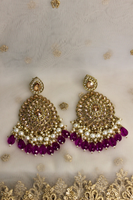 This image displays a pair of elegant Ishita earrings, positioned against a backdrop embellished with golden lace. The earrings are crafted with a detailed golden structure, adorned with sparkling gemstones and delicate pearl accents.